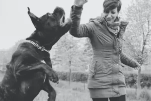 Woman in a hooded coat high-fives a large dog in a park; both appear joyful in a black-and-white scene.