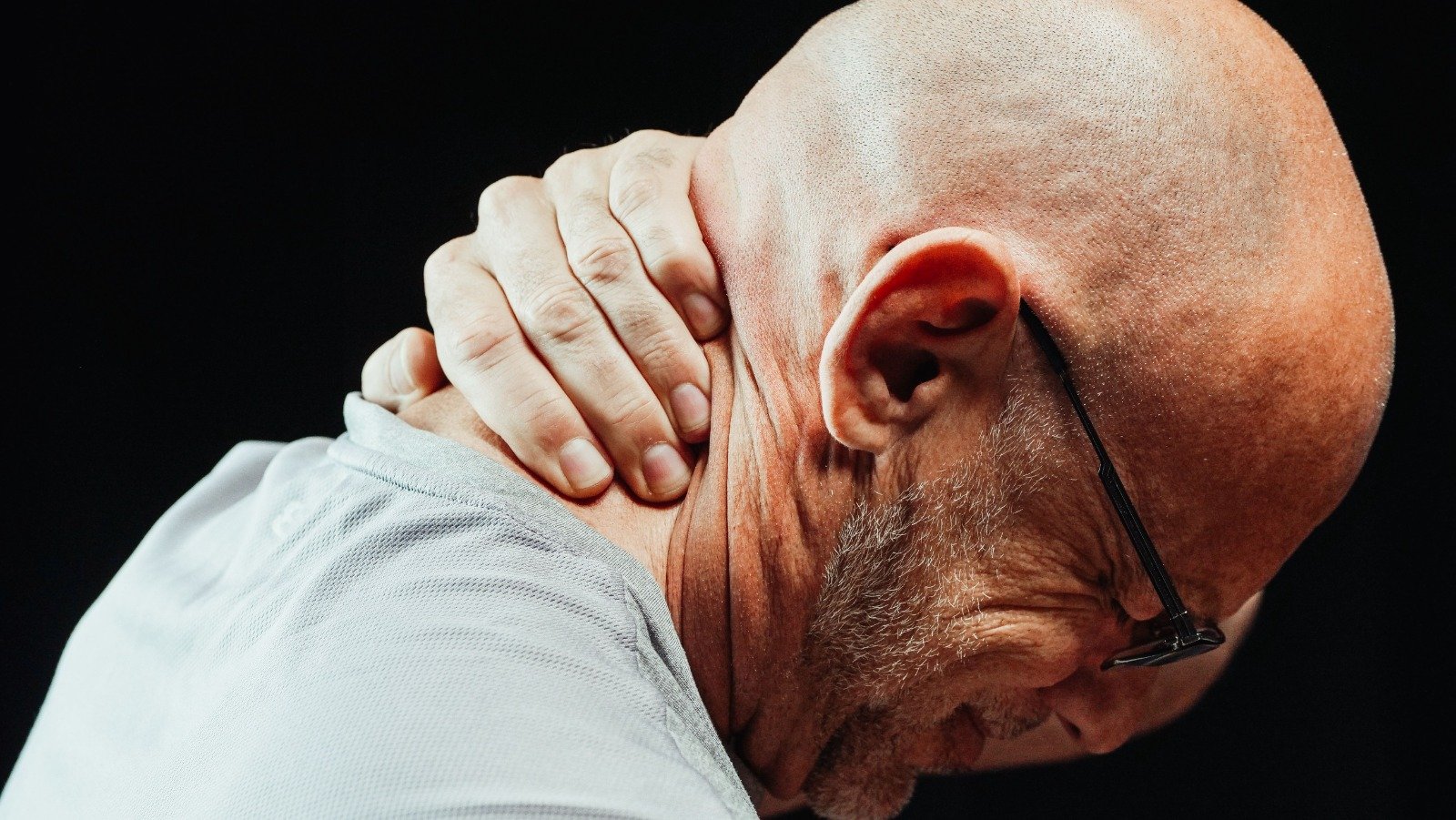 Man with glasses touching his neck in apparent pain, head tilted downward against a dark background