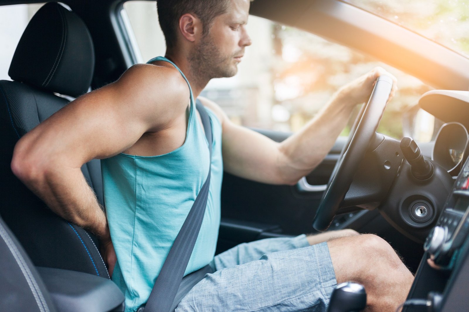 Man driving a car in a turquoise sleeveless shirt, gripping the steering wheel with a seatbelt on.