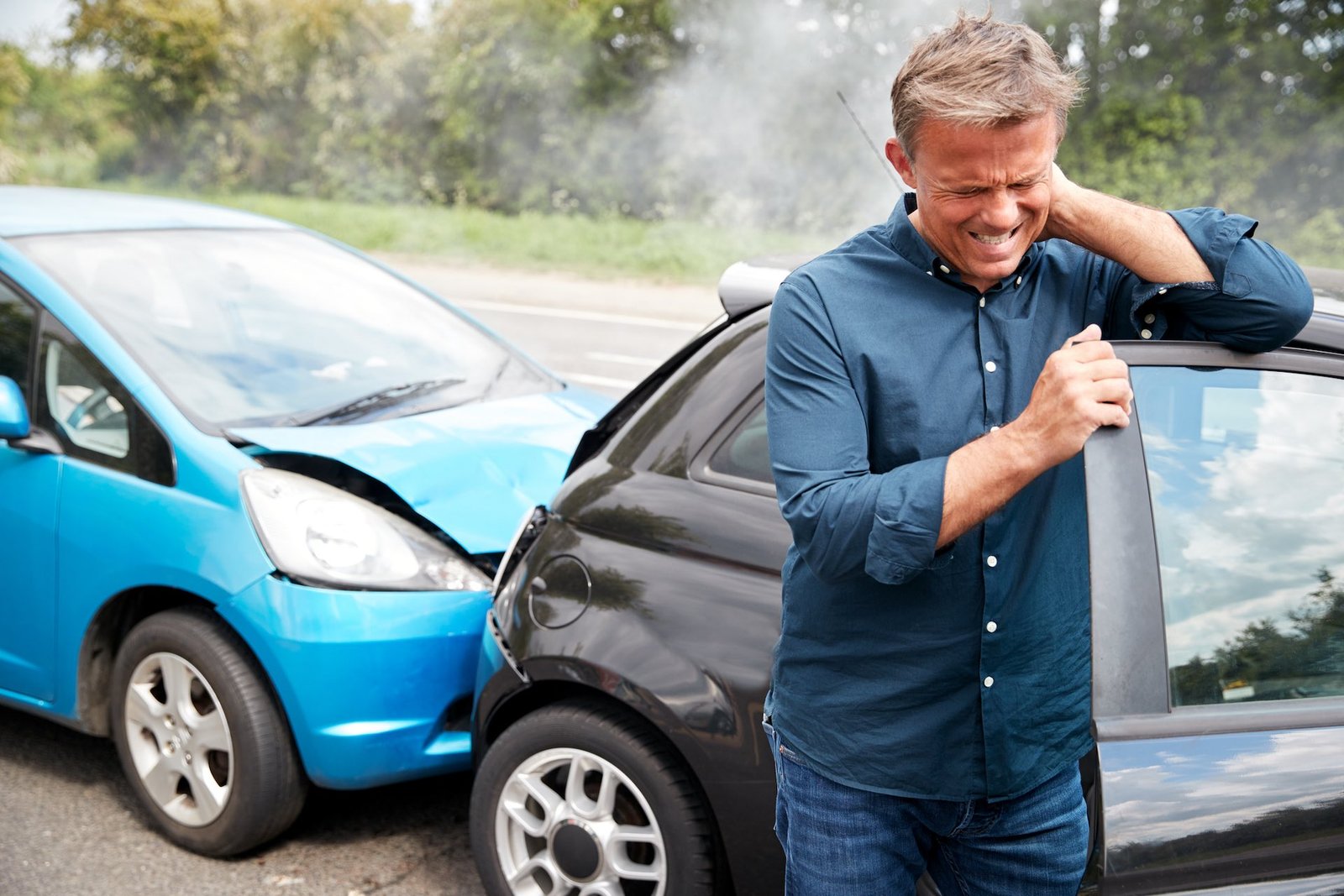 Man in a blue shirt winces in pain, leaning on a black car with its door open after a crash; a damaged blue car is nearby in a roadside setting.